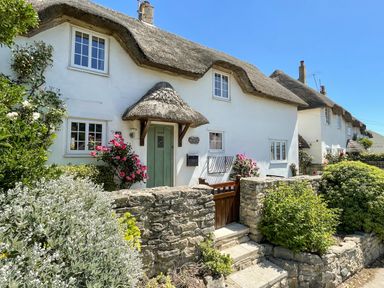 A thatched roof cottage with a green door surrounded by stone walls and flowering bushes at Cajun Cottage in West Lulworth