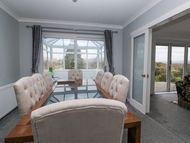 A dining room with a glass table and beige upholstered chairs next to a conservatory at Eildon Lodge in Gosforth