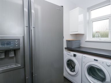 A utility room with a silver fridge freezer a washing machine and a dryer under a black countertop and a white cabinet on the wall at Eildon Lodge in Gosforth