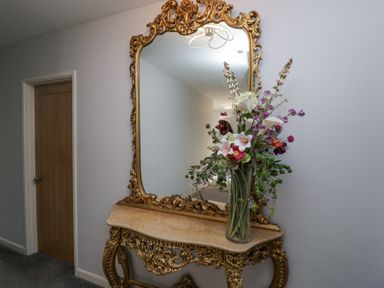 A hallway with an ornate gold framed mirror above a matching console table and a vase of flowers at Eildon Lodge in Gosforth