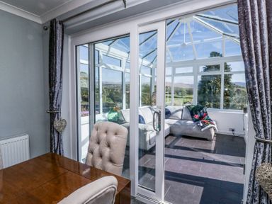 A dining area with upholstered chairs and wooden table next to a glass door leading to a conservatory with a sectional sofa and large windows at Eildon Lodge in Gosforth