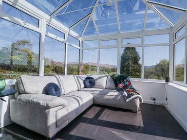 A conservatory with a grey sectional sofa two round blue pillows and a patterned blanket at Eildon Lodge in Gosforth