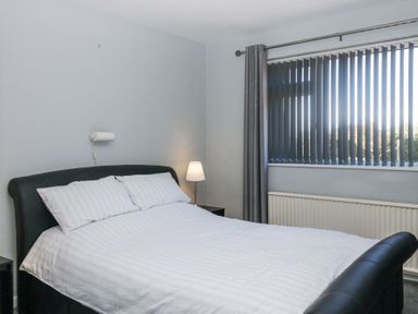 A bedroom with a double bed with white bedding a bedside table with a lamp and a window with vertical blinds and curtains at Eildon Lodge in Gosforth