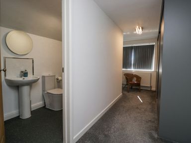 A hallway with carpet leading to a room with a wicker chair and a bathroom with a toilet and sink at Eildon Lodge in Gosforth