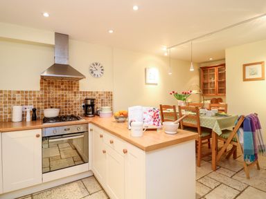 A kitchen with a stove coffee maker and fruit bowl next to a dining table with chairs and flowers at Fox Hat Cottage in Chagford