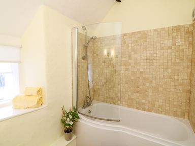 A bathroom with a bathtub shower combination tiled walls glass shower screen rolled towels on a window ledge and a plant at Fox Hat Cottage in Chagford