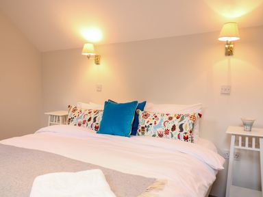 A bedroom with a double bed white bedding patterned cushions blue cushions two wall lamps and two bedside tables at Fox Hat Cottage in Chagford