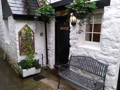 A black door with the number 5 and a bench in front of a white stone wall with a window and hanging plants at Fox Hat Cottage in Chagford