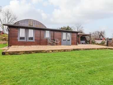A wooden cabin with gray window frames and doors on a gravel patio with chairs and table on a lawn at Westfield Lodge in Terrington near Sheriff Hutton