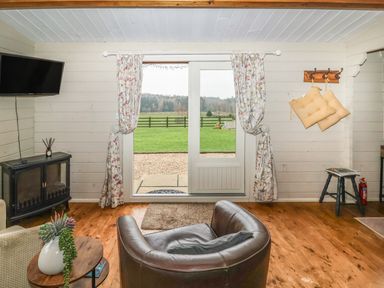 A living room with a leather chair a round wooden table with a plant floral curtains over glass doors opening to a green field at Westfield Lodge in Terrington near Sheriff Hutton