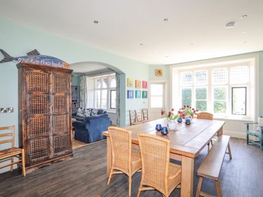 A dining room with a wooden table surrounded by wicker chairs and bench with flowers on the table at Bryn Henllan in Criccieth