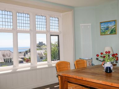 A dining area with a wooden table and wicker chairs next to large leaded windows showing a view of rooftops and a castle at Bryn Henllan in Criccieth