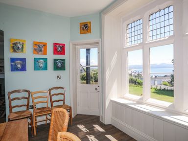 A room with wooden chairs and colorful animal paintings on the wall next to a window with a view of a garden and distant hills at Bryn Henllan in Criccieth
