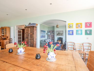A dining room with a wooden table with two vases of red flowers and chairs with an open view to a living room with sofas a fireplace and wall art at Bryn Henllan in Criccieth