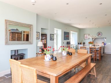 A dining area with a wooden table, wicker chairs, and red flowers in vases next to a kitchen with stools at Bryn Henllan in Criccieth