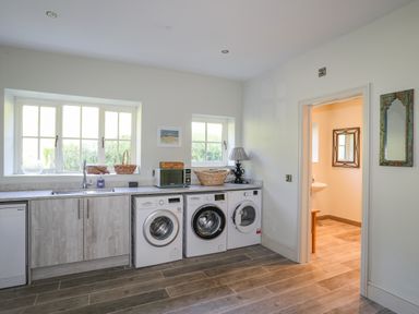 A laundry room with three washing machines a sink microwave lamp and baskets on the counter at Bryn Henllan in Criccieth