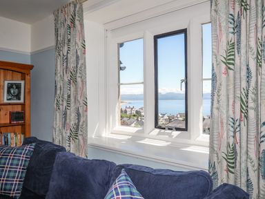 A living room with a blue sofa and patterned curtains by a window overlooking a coastal town at Bryn Henllan in Criccieth