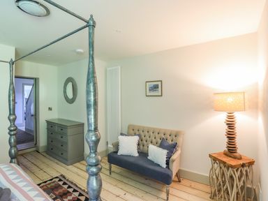 A bedroom with a four-poster bed, a small sofa with cushions, a lamp on a wooden side table, a chest of drawers, and a round mirror on the wall at Bryn Henllan in Criccieth