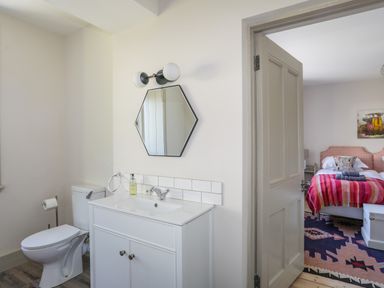 A bathroom with a toilet, white vanity with sink and hexagonal mirror next to a door opening to a bedroom with a bed, colorful blanket and patterned rug at Bryn Henllan in Criccieth