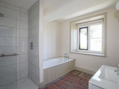 A bathroom with a shower area with tiled walls a bathtub below a window a sink and a rug on the floor at Bryn Henllan in Criccieth