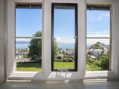 View through three windows of houses trees green lawn and a castle on a hill by the sea at Bryn Henllan in Criccieth