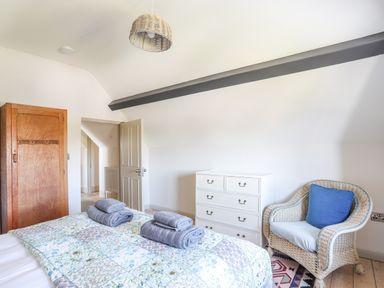 A bedroom with a bed covered by a patterned quilt and towels on top a wooden wardrobe a white dresser and a wicker chair with a blue cushion at Bryn Henllan in Criccieth