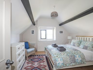 A bedroom with a double bed covered with a patchwork quilt two rolled towels on the bed a wicker chair with a cushion a chest of drawers and a window at Bryn Henllan in Criccieth