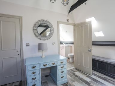 A room with a light blue desk, a lamp, a round mirror on the wall, and a door open to a bathroom with a toilet at Bryn Henllan Criccieth