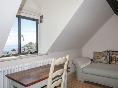 A room with a window showing a view of a hill and castle a wooden table with a chair and a sofa with cushions at Bryn Henllan in Criccieth