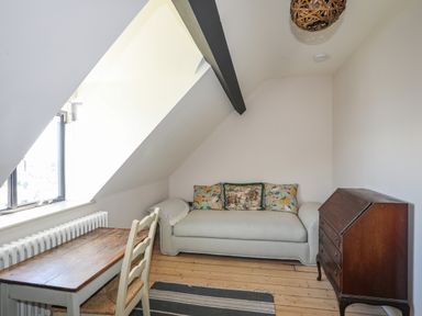 A small room with a slanted ceiling containing a wooden table with chairs a light-colored sofa with cushions a wooden chest of drawers and a window at Bryn Henllan in Criccieth