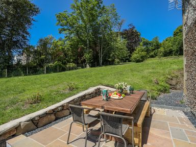 An outdoor patio area with a wooden table and chairs next to a grassy slope with trees at Bryn Henllan in Criccieth