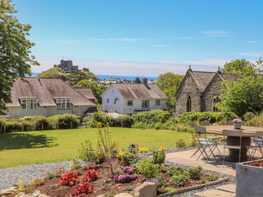 A garden with flowers and a patio table with chairs overlooking houses a stone building and a castle on a hill in Criccieth