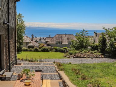 A backyard with a wooden table and chairs on a patio overlooking houses trees and the sea at Bryn Henllan in Criccieth