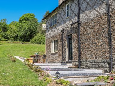 A patio area with a table and chairs beside a house with pebble dash walls and a grassy garden at Bryn Henllan in Criccieth