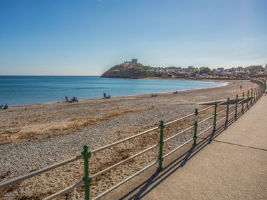 A pebble and sand beach with a railing along a promenade and a castle on a hill overlooking the sea at Bryn Henllan in Criccieth