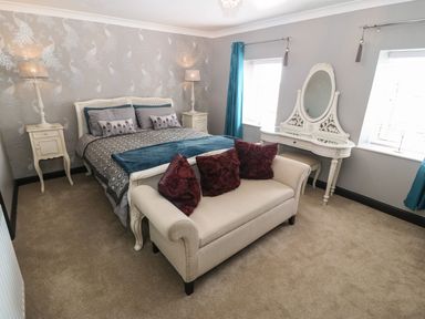 A bedroom with a double bed and two bedside tables with lamps a white vanity desk and a beige loveseat with red pillows at Trewan House Bryngwran near Rhosneigr Rhosneigr