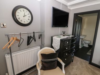 A room with a wall clock a clothes rack with hangers an upholstered chair a black chest of drawers with a white runner and a wall-mounted television at Trewan House Bryngwran near Rhosneigr Rhosneigr