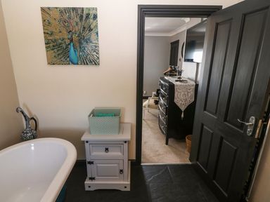A bathroom with a white bathtub a small cabinet with a basket and a peacock painting on the wall looking into a bedroom with a black dresser and a wall-mounted TV at Trewan House Bryngwran near Rhosneigr Rhosneigr