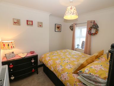 A bedroom with a bed covered in yellow floral bedding a black chest of drawers with a lamp and decorative items and a window with pink curtains at Trewan House Bryngwran near Rhosneigr Rhosneigr