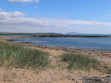 A sandy beach with grass and pebbles by the sea with hills in the background at Trewan Lodge in Bryngwran near Rhosneigr