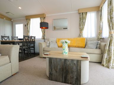 A living room with a beige sofa a yellow throw a wooden coffee table with a vase of flowers and a dining table with chairs at Trewan Lodge in Bryngwran near Rhosneigr