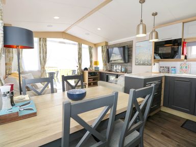 A dining table with chairs next to a kitchen and a living area with a sofa and television at Trewan Lodge in Bryngwran near Rhosneigr