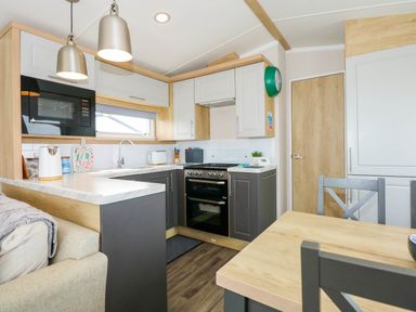 A kitchen with wooden cabinets grey lower cupboards a stove a microwave and a wooden dining table with grey chairs at Trewan Lodge in Bryngwran near Rhosneigr