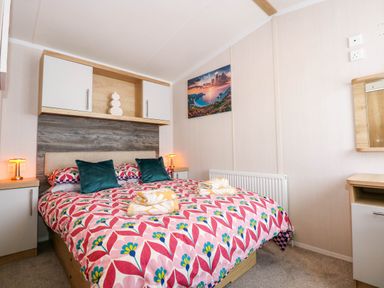 A bedroom with a bed and floral bedding two bedside tables with lamps cabinets above the bed and a wall picture at Trewan Lodge in Bryngwran near Rhosneigr