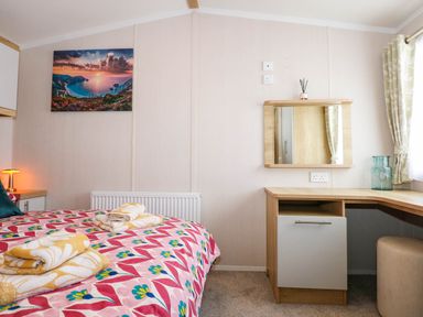 A bedroom with a bed, a wooden dressing table with a mirror, and a window with patterned curtains at Trewan Lodge in Bryngwran near Rhosneigr