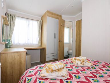 A bedroom with a bed covered in a patterned bedspread with folded towels on top a wooden wardrobe and desk near a window with curtains at Trewan Lodge in Bryngwran near Rhosneigr