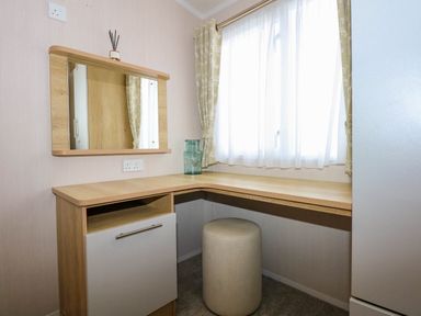A corner desk with a mirror above a drawer and a cylindrical stool under a window with curtains at Trewan Lodge in Bryngwran near Rhosneigr