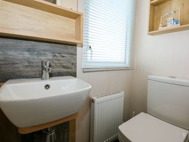 A bathroom with a white sink, toilet, radiator, window with blinds and wooden shelves at Trewan Lodge Bryngwran near Rhosneigr