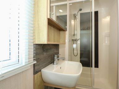 A bathroom with a white sink and faucet a wooden cabinet a shower with a glass door and a window with blinds at Trewan Lodge Bryngwran near Rhosneigr