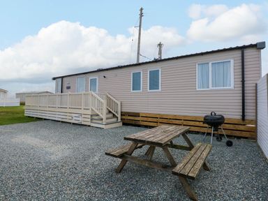 An exterior view of a single-story mobile home with a small deck and stairs a wooden picnic table and a barbecue grill on a gravel surface at Trewan Lodge in Bryngwran near Rhosneigr